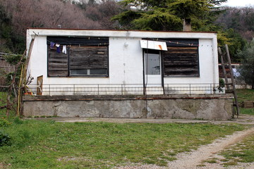 Small dilapidated house without roof with broken wooden blinds on windows, improvised cloth line, broken concrete terrace and uncut grass in front and dense winter forest vegetation in background