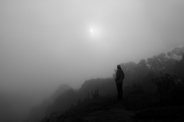 Black and white photo of man standing on the peak of sandstone. Misty valley. Foggy mountains. Sun in the fog.