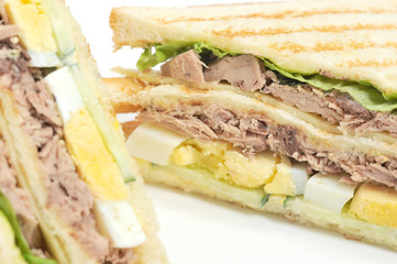 Sandwich of white bread triangular shape on white background. The filling of the sandwich consists of pieces of tuna, hard boiled eggs, lettuce leaves, cheese and sauce. Close-up. Macro photography.