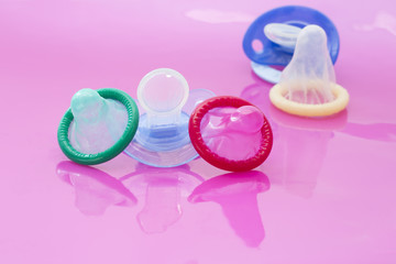 Contraception concept. Condoms green, red and white and blue pacifiers on pink background