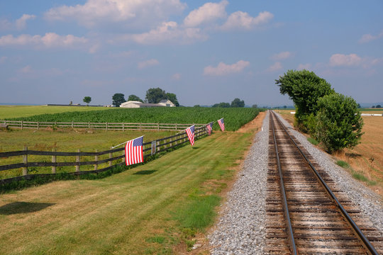 American flags on a fence line running parallel to the railroad tracks with a big blue sky and clouds
