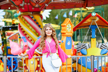 Bright summer concept. A cheerful blonde girl in pink jacket is having fun in the amusement Park. The woman looks happily at the camera and posing in front of the children's carousel with cotton candy