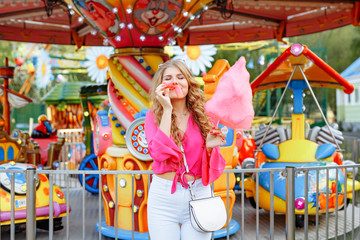 Bright summer concept. A cheerful blonde girl in pink jacket is having fun in the amusement Park. The woman looks happily at the camera and posing in front of the children's carousel with cotton candy
