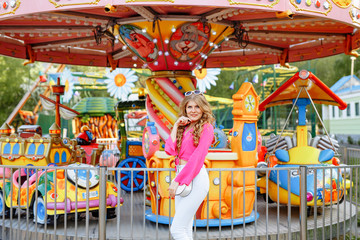 Fototapeta na wymiar Bright summer concept. A cheerful blonde girl in pink jacket and stylish sunglasses is having fun in the amusement Park. Cheerful woman smiling against bright carousel