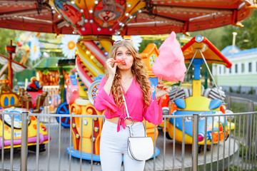 Obraz na płótnie Canvas Bright summer concept. A cheerful blonde girl in pink jacket is having fun in the amusement Park. The woman looks happily at the camera and posing in front of the children's carousel with cotton candy