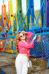 Bright summer concept. A cheerful blonde girl in pink jacket and stylish sunglasses is having fun in the amusement Park. Cheerful woman smiling against bright carousel