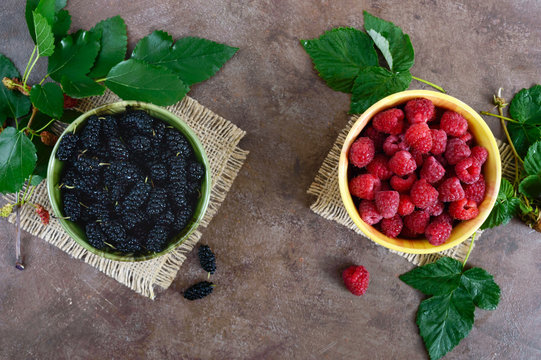 Summer berries: ripe black mulberry and juicy raspberries in bowls, scattered leaves on a vintage background. Harvest from the garden. Top view.