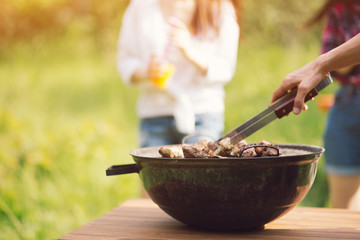 Big grill of tasty meat sitting on wooden outdoor table. Person taking some meat out of round metal bowl using tongs.