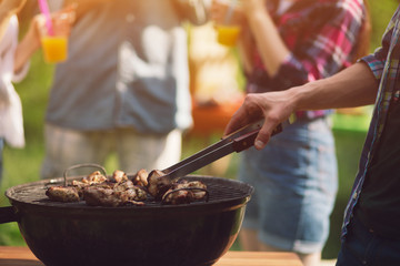 Tasty meat getting fried on grill. Person wearing button up shirt flipping meat over on fry grill otside in park. Group of people blurred in background.