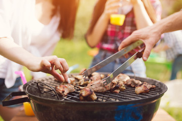 People taking meet of grill. Hand of woman picking piece of frying meat with her hands next to man fliping it over with kitchen tongs.
