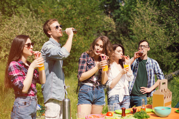 Fototapeta premium Five people outdoors on sunny day eating and drinking. Joyful company of young people out on picnic having drinks and food. Trees in background.
