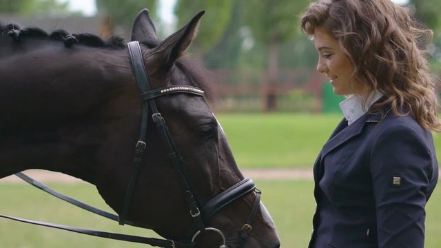 a young girl jockey is standing next to a brown racehorse in a harness and gently stroking her. the girl hugs the horse on her head. . juicy selenium grass in the background. the rider looks into the