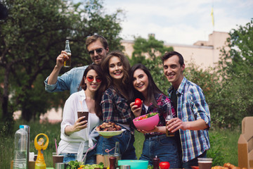 Cheerful young people smiling looking happy. Friends dressed up in light summer clothes taking group picture with food and drinks on picnic.