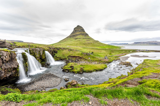 Landscape view of famous Kirkjufell Mountain and Waterfall landmark with long exposure smooth water wide angle with people tourists, cars in parking lot green overcast cloudy day