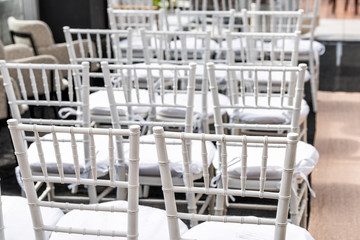 White wedding chairs closeup for ceremony with background of rows of many seats pattern, aisle and podium in venue, restaurant, building outdoors
