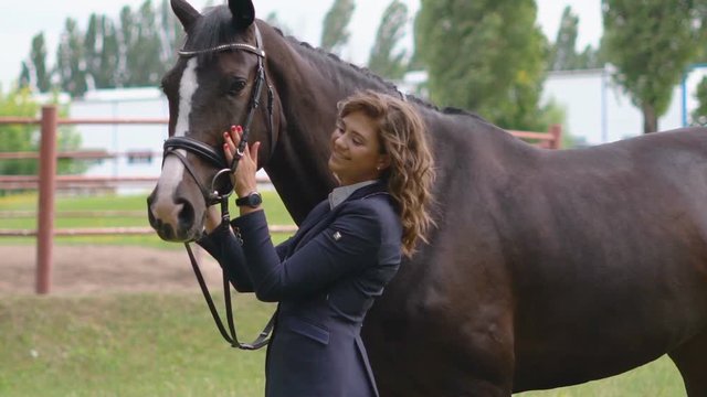 a young girl jockey is standing next to a brown racehorse in a harness and gently stroking her. the girl takes the horse on her head. . juicy selenium grass in the background. HD slow motion