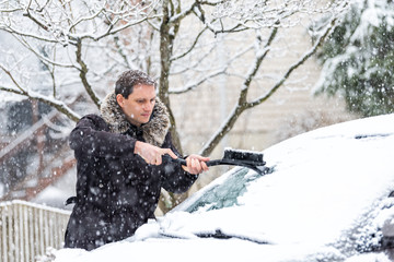 Young man cleaning car windshield from snow, ice with brush and scraper tool during snowfall during winter snowing snowflakes falling - obrazy, fototapety, plakaty