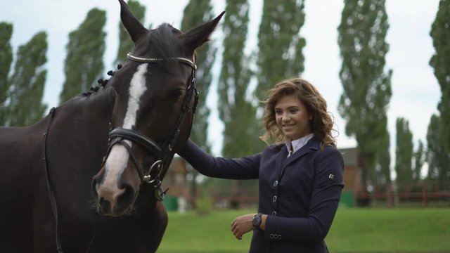a young girl jockey is standing next to a brown racehorse in a harness and gently stroking her. the girl takes the horse on her head. . juicy selenium grass in the background. 4K