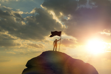 silhouette of a man on top of a mountain holding a flag of victory, against the sky and sunset....