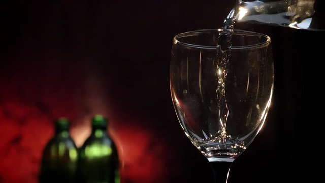 Pouring white wine into glass shooting, two bottles and red background, alcohol drink filling goblet