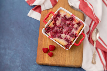 Homemade Raspberry Cobbler in Red Cookware on Wooden Cutting Board beside a Red & White Kitchen Towel on a Blue Background