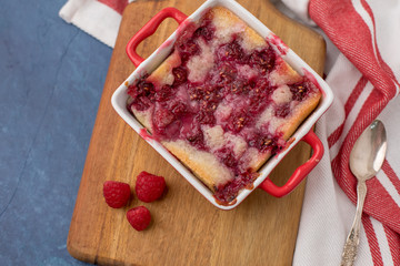 Homemade Raspberry Cobbler in Red Cookware on Wooden Cutting Board beside a Red & White Kitchen Towel on a Blue Background