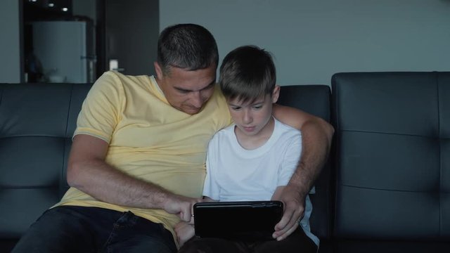 Father and son are sitting on the sofa watching movies and playing games on the tablet. A happy family
