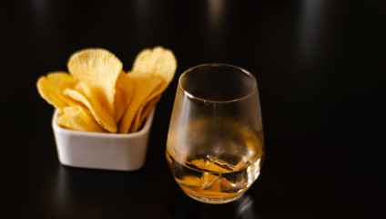 glass of whiskey with ice cubes and salty snacks on a black background
