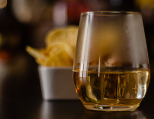 glass of whiskey with ice cubes and salty snacks on the background of bottles and bar