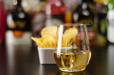 glass of whiskey with ice cubes and salty snacks on the background of bottles and bar