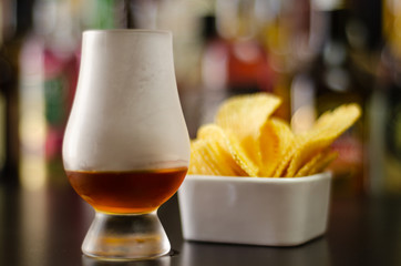 glass of whiskey with ice cubes and salty snacks on the background of bottles and bar