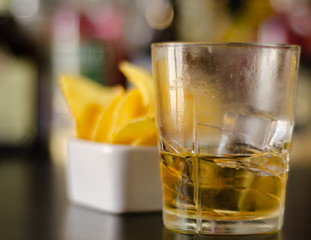 glass of whiskey with ice cubes and salty snacks on the background of bottles and bar