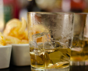 glass of whiskey with ice cubes and salty snacks on the background of bottles and bar