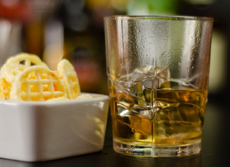 glass of whiskey with ice cubes and salty snacks on the background of bottles and bar