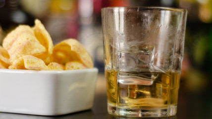 glass of whiskey with ice cubes and salty snacks on the background of bottles and bar