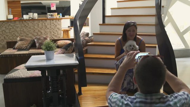 A man takes pictures of a woman sitting on the stairs in the house with a small dog in her arms