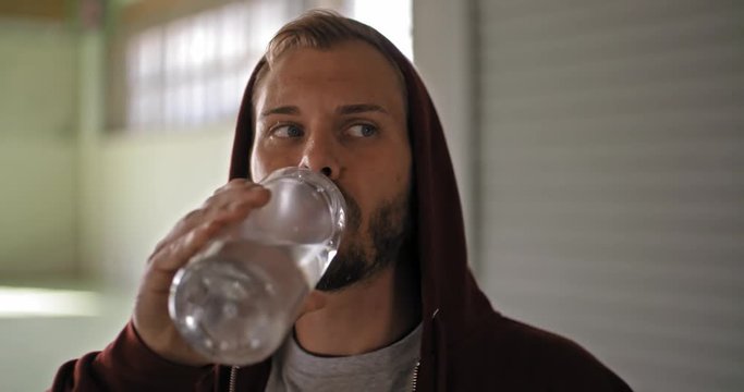 Young adult man with hooded sweatshirt drinking water resting during fitness sport workout .Grunge industrial urban training.4k slow motion video