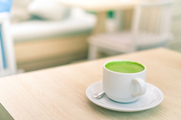 A white cup of green tea latte on wood table in with blur coffee cafe shop and nature green.