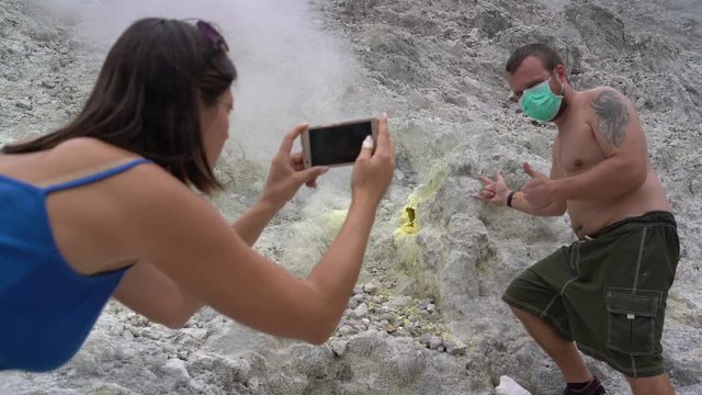 A woman is taking pictures of a man next to a fumarole on a smartphone