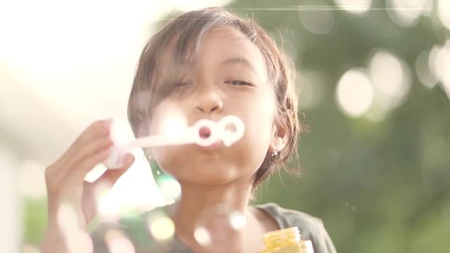 Happy toothless Asian little girl blowing soap bubble at the park with blurred nature background