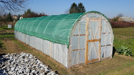 Plastic greenhouse with wooden doors, thick green net on top situated in garden surrounded with plants and uncut grass with houses and blue sky in background