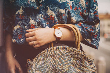 street style fashion details. woman wearing a summer shirt and a white and brown analog wrist watch, holding a round straw purse. perfect summer fashion accessories. warm vintage grade - obrazy, fototapety, plakaty
