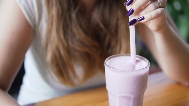 Young beautiful european girl mixing and drinks cold milk cocktail with blueberry in a cafe in hot summer day. Women drinking smoothies throught the straw.