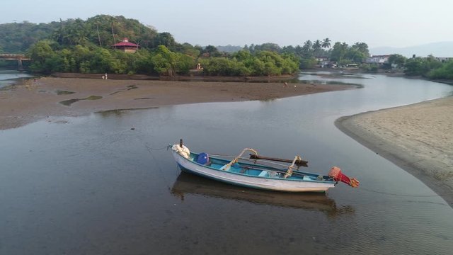 Aerial view. Flying around white-blue Indian traditional wooden boat with side float anchored on a shallow river at sunset. Fishing nets near on the shore. 