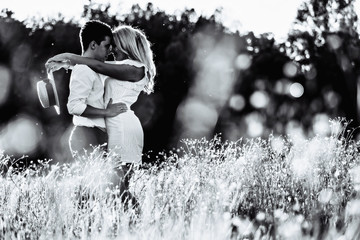 Awsome happy couple have fun in a field sunset background. Handsome Caucasian guy wearing white shirt gentle hugs a smiling girl with blond hair in white dress. Lifestyle and travel concept.