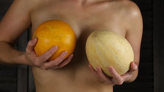 Young happy woman holds two melons above her breast. Plastic surgery and silicone implants concept. slow motion