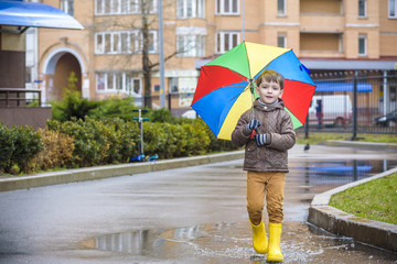 Little boy playing in rainy summer park. Child with colorful rainbow umbrella, waterproof coat and boots jumping in puddle and mud in the rain. Kid walking in autumn shower Outdoor fun by any weather