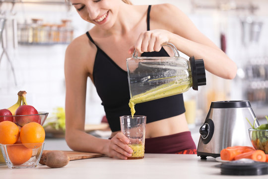 Young woman pouring tasty healthy smoothie into glass at table in kitchen