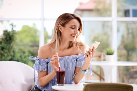 Young woman using mobile phone while drinking tasty healthy smoothie at table, indoors
