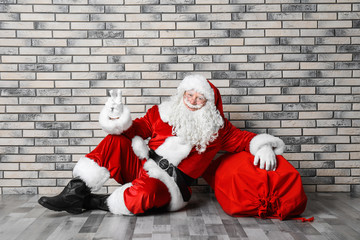 Authentic Santa Claus with big red bag full of gifts sitting on floor near brick wall